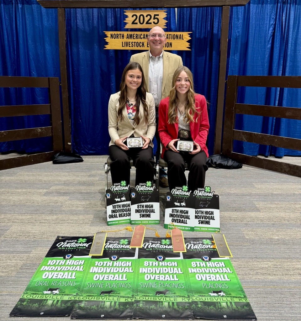 Two girls seated with a number of awards in front of the 2025 North American International Livestock Expedition sign. Behind them stands a man in a blazer.